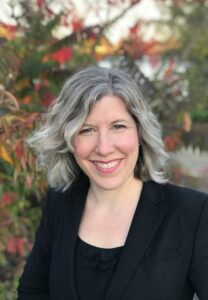 woman with gray hair wearing a black shirt and jacket smiling for the camera with trees behind her