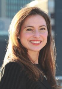 woman with brown hair wearing a black top smiling for camera with buildings in the background